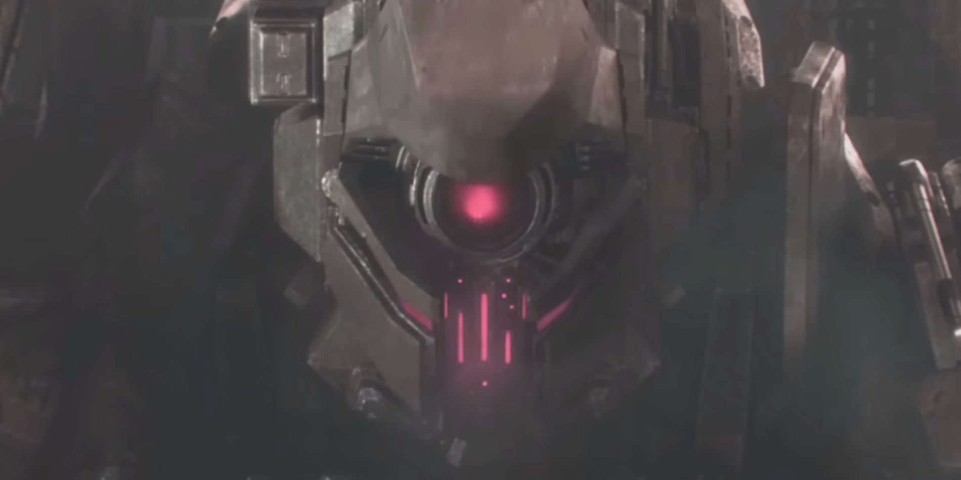 Uma imagem aproximada de uma grande cabeça de mech Armored Core cinza é vista com luzes vermelhas brilhantes saindo de seu rosto, voltada diretamente para o observador.