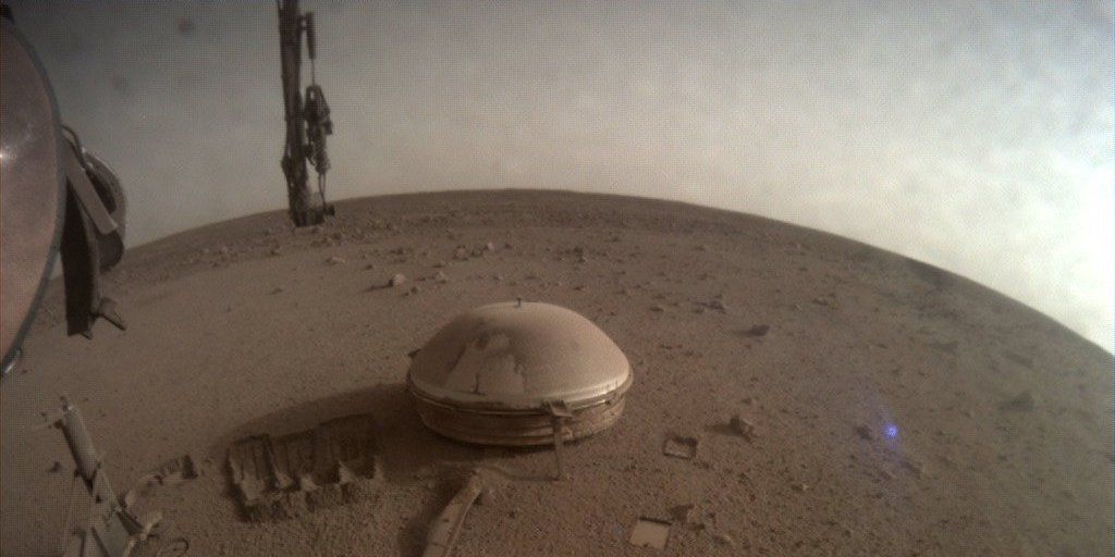 The InSight lander's final photo, showing the dusty brown Mars landscape and a light gray sky, with the dome of its seismometer showing