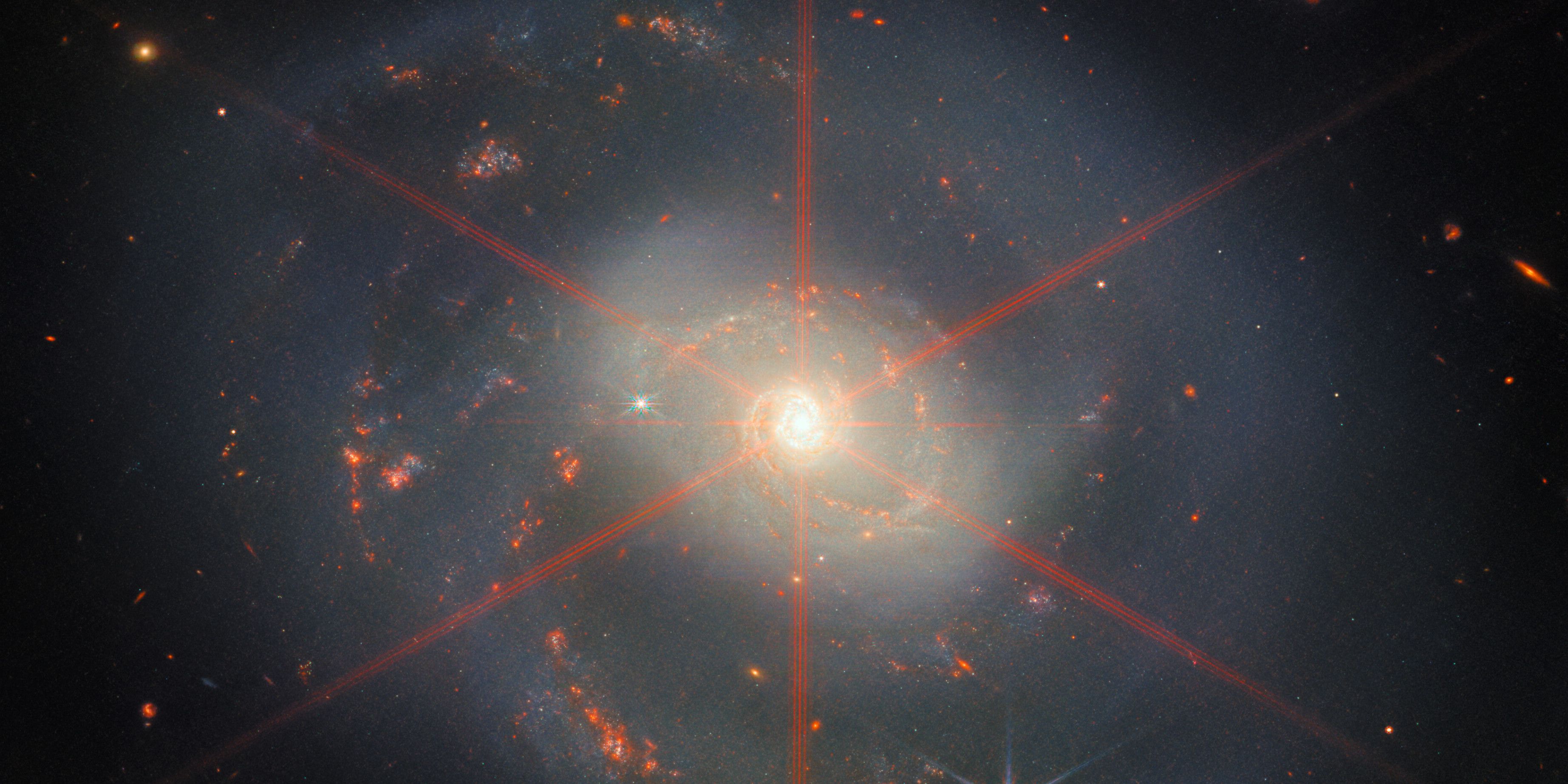 An image from the James Webb Space Telescope showing a bright, spiral galaxy with six red beams emanating from its center