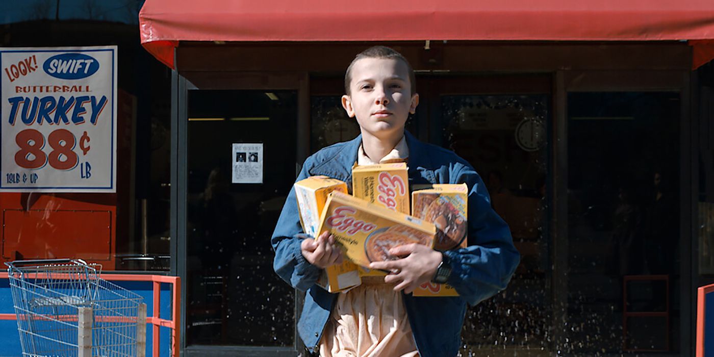 Onze de Stranger Things em frente a uma loja, portas de vidro quebradas segurando várias caixas de Eggos.