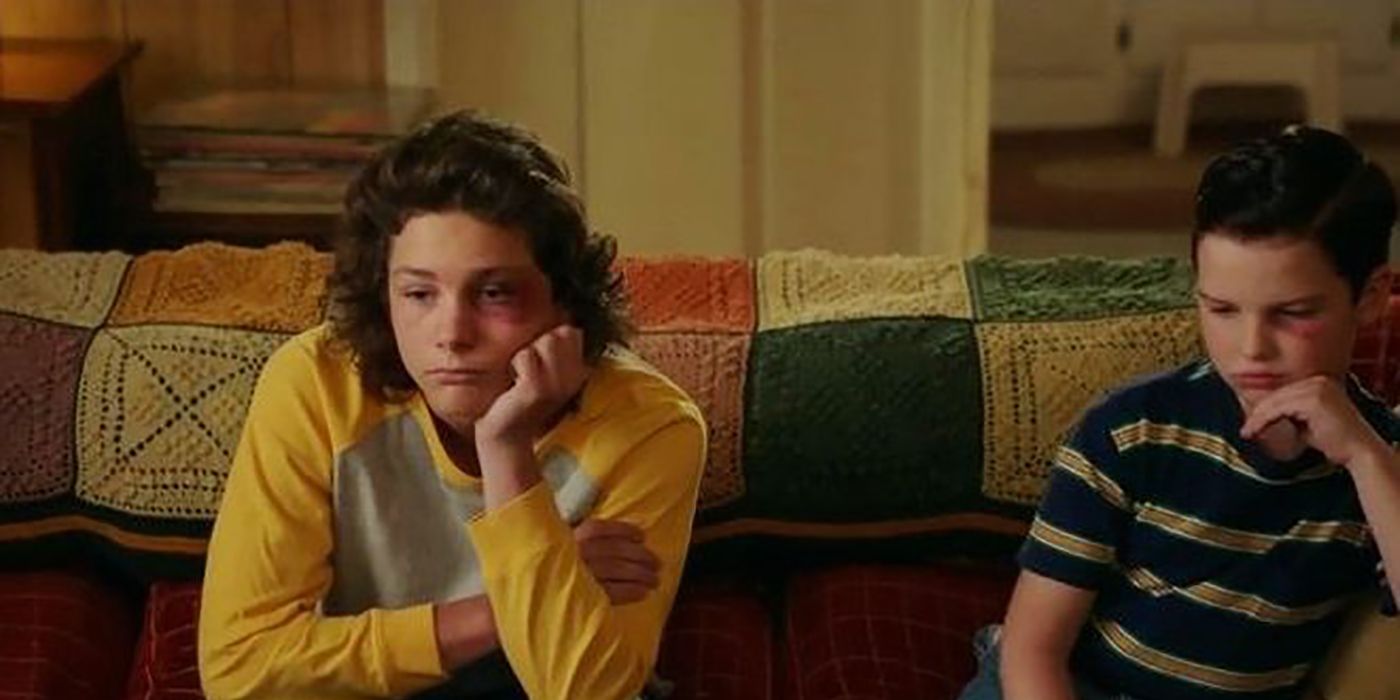 Georgie from Young Sheldon sitting on the couch with Sheldon, both with black eyes and looking dejected.