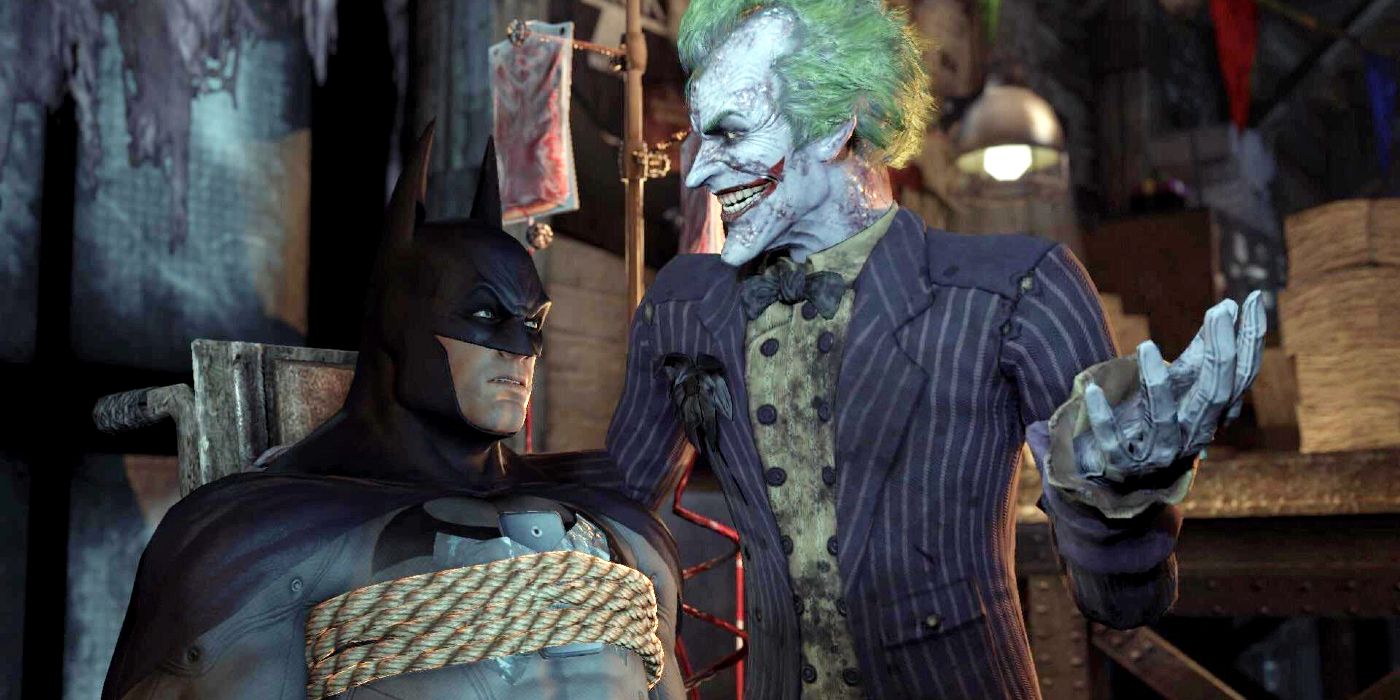 Screenshot of a cutscene from Batman: Arkham City. Batman is tied up on a chair scowling at Joker, who has one hand planted tauntingly on the Dark Knight's left shoulder.