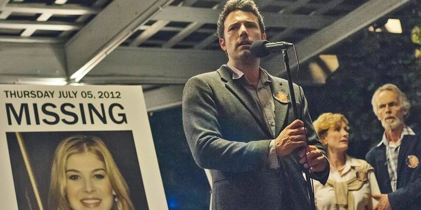 Nick (Ben Affleck) standing at a microphone next to a missing Amy poster in Gone Girl