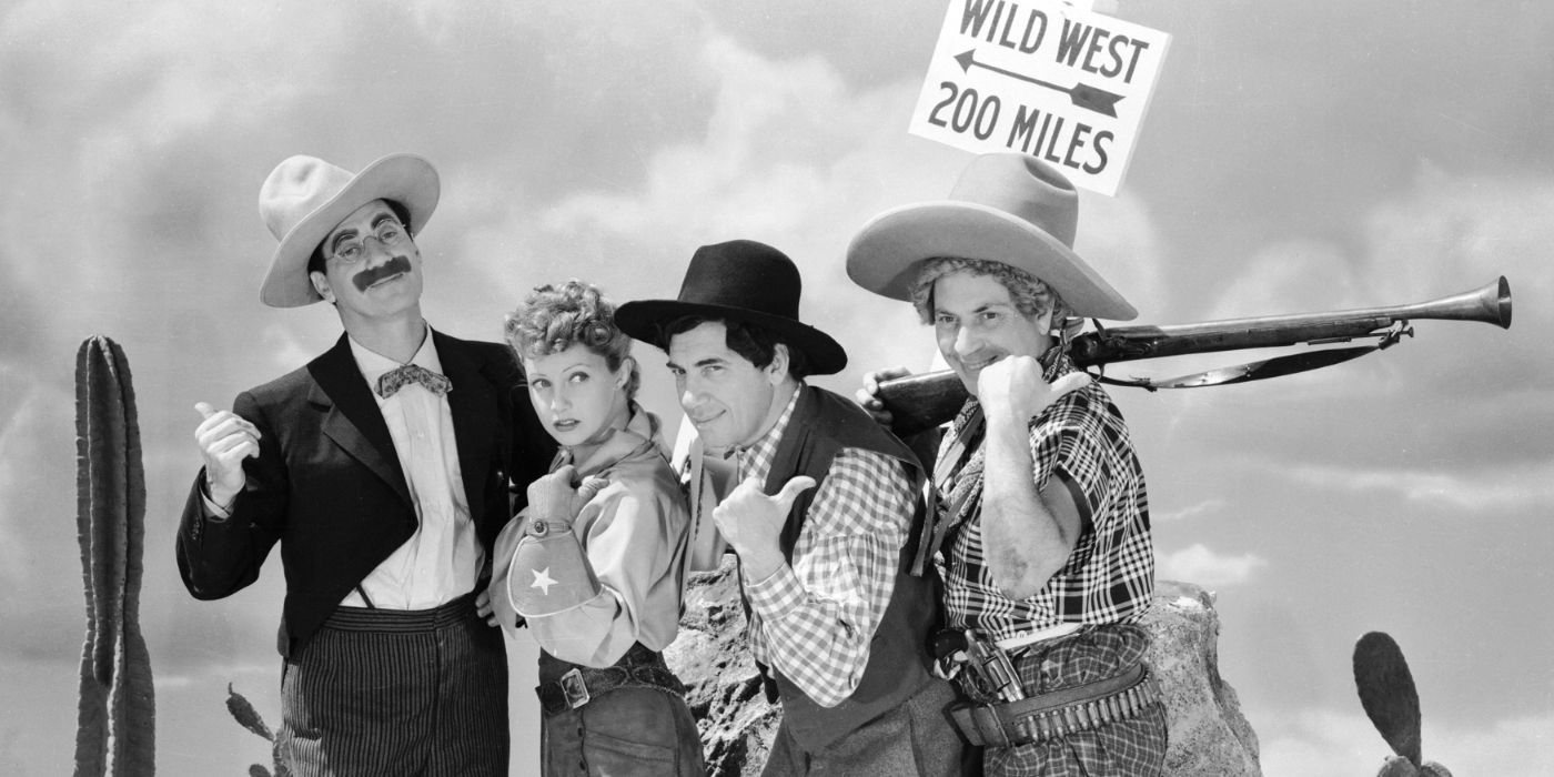 groucho chico and zippo marx and diana lewis standing in a line in cowboy gear with their thumbs sticking out in go west, a sign with "wild west 200 miles" sits behind them