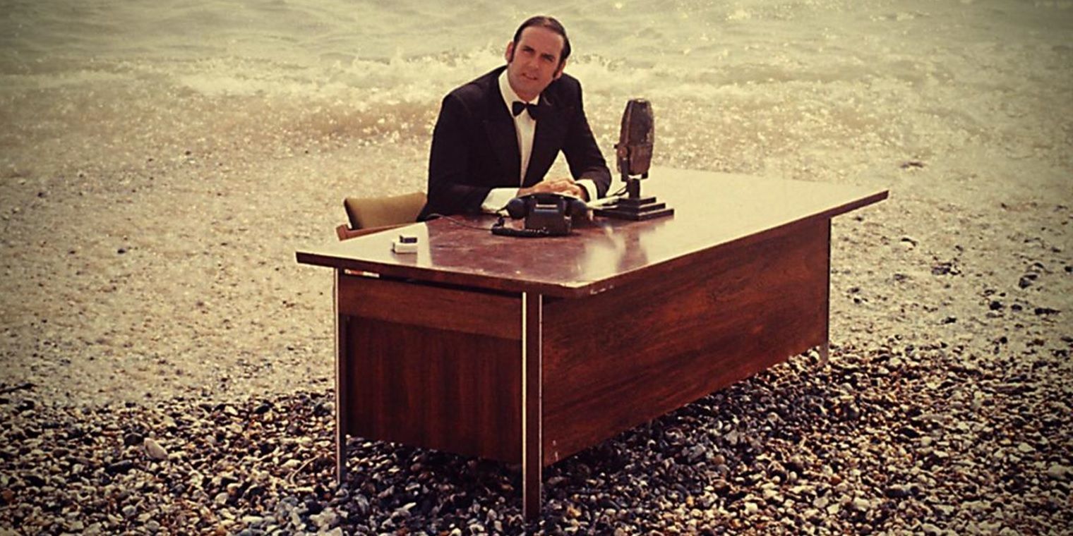 John Cleese sits at a desk on the beach in And Now for Something Completely Different