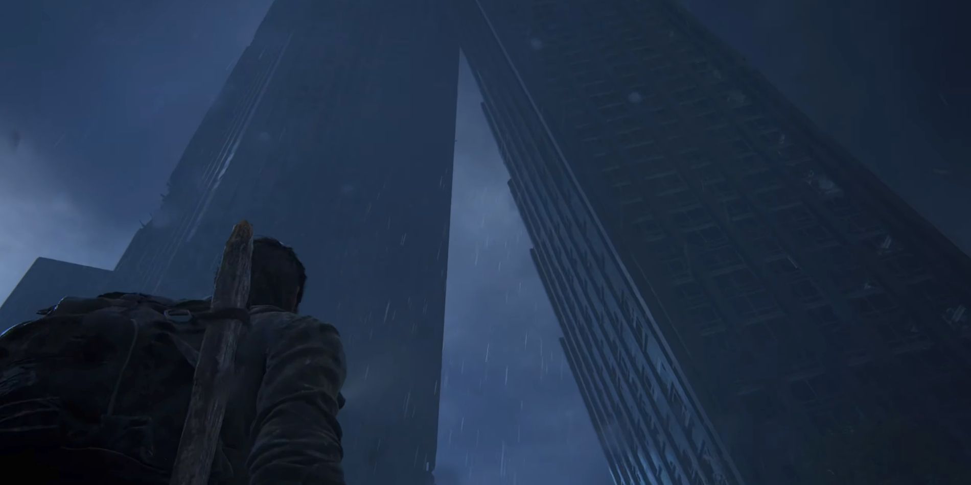Joel from The Last of Us Part 1 looking up at two large skyscrapers, one of which is broken and leaning on the side of the other.