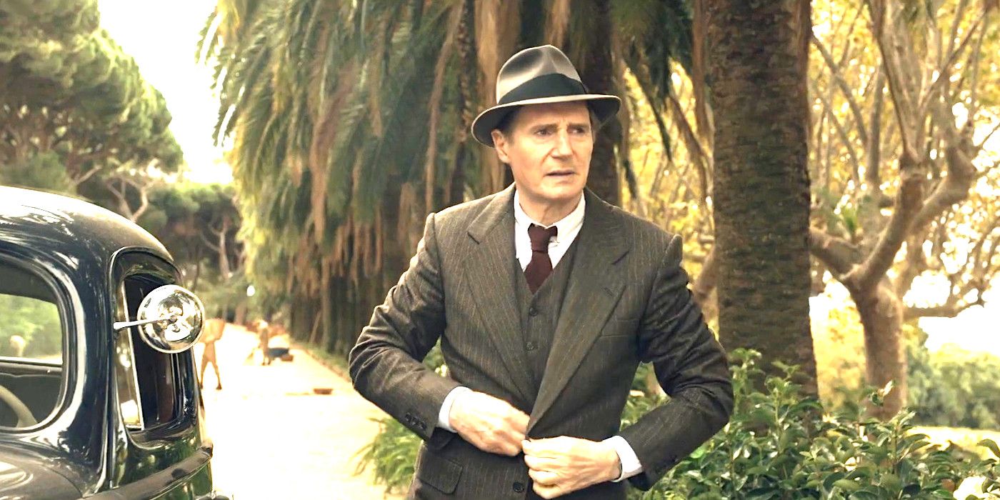 Liam Neeson in the movie Marlowe wearing a suit and hat, in the middle of buttoning his suit jacket while walking away from his 1930s car on a road lined with palm trees