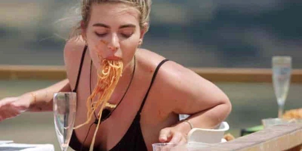 Uma garota em um minúsculo maiô preto come um espaguete desleixado na Ilha do Amor.
