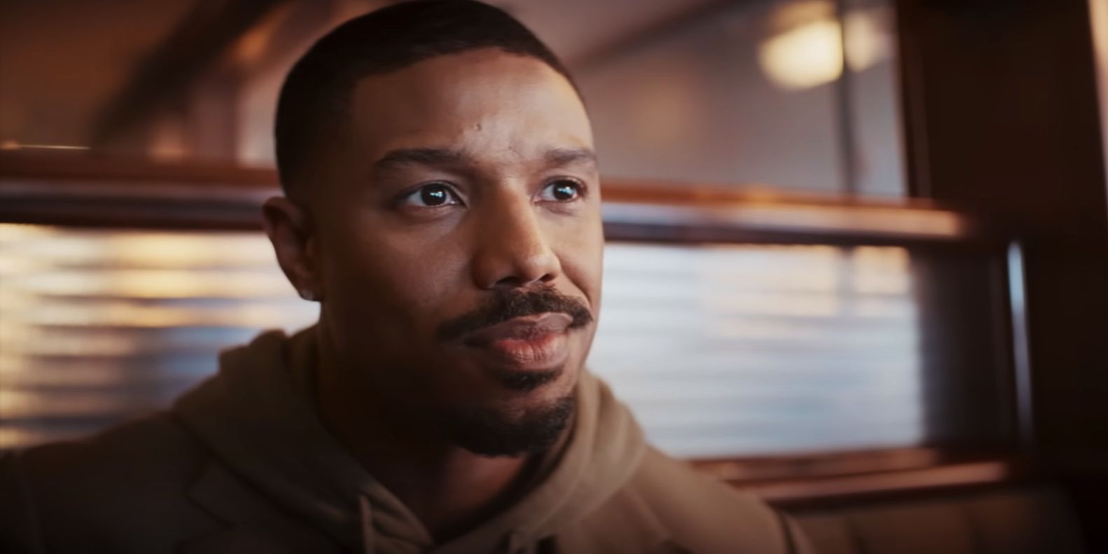 Michael B Jordan as Adonis Creed sitting in booth smiling slightly in Creed 3