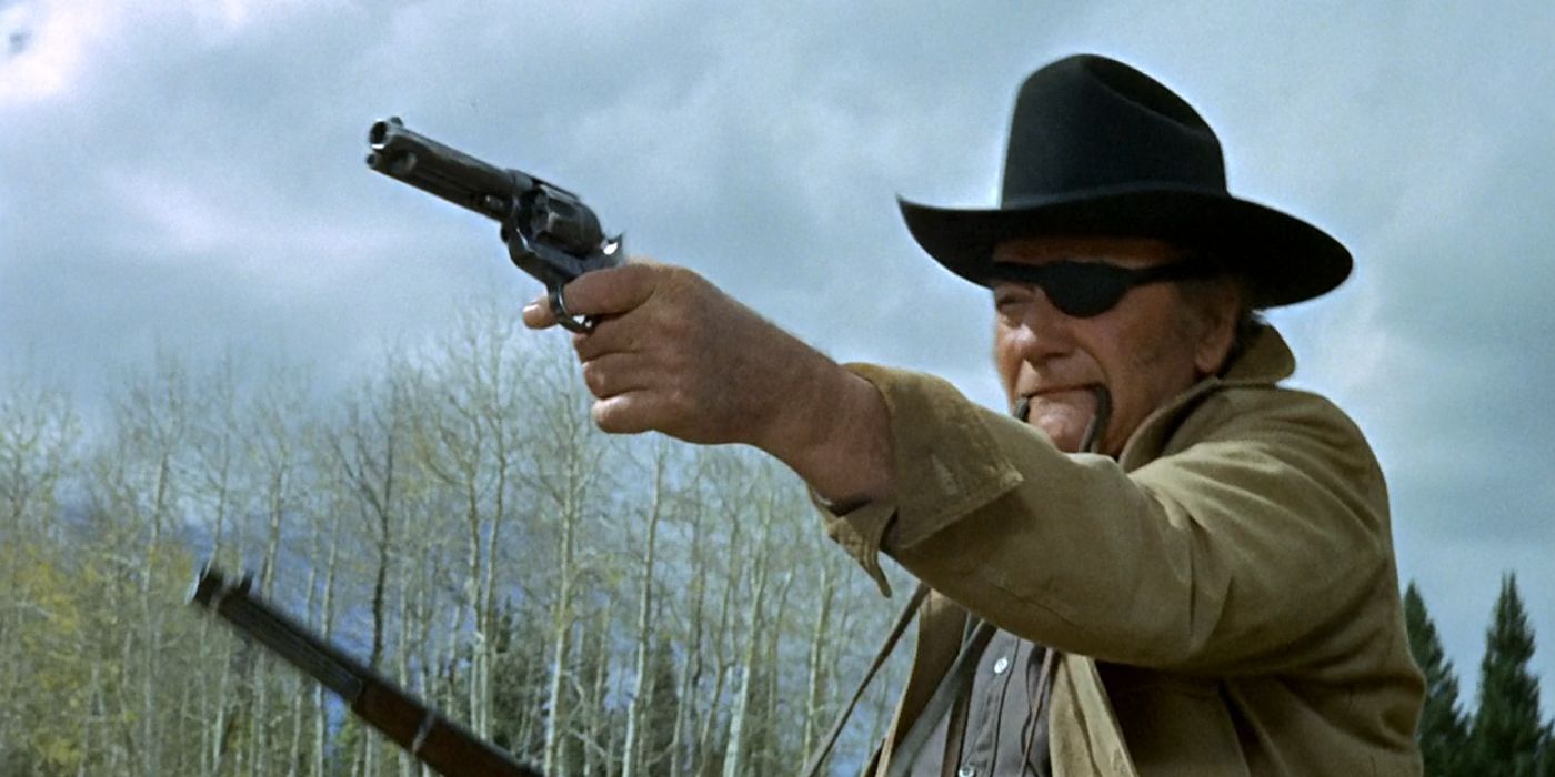 Image of John Wayne's Rooster Cogburn in True Grit (1969). Shot shows Rooster wielding both a revolver and a repeating rifle on horseback, firing it off to the left of the camera.