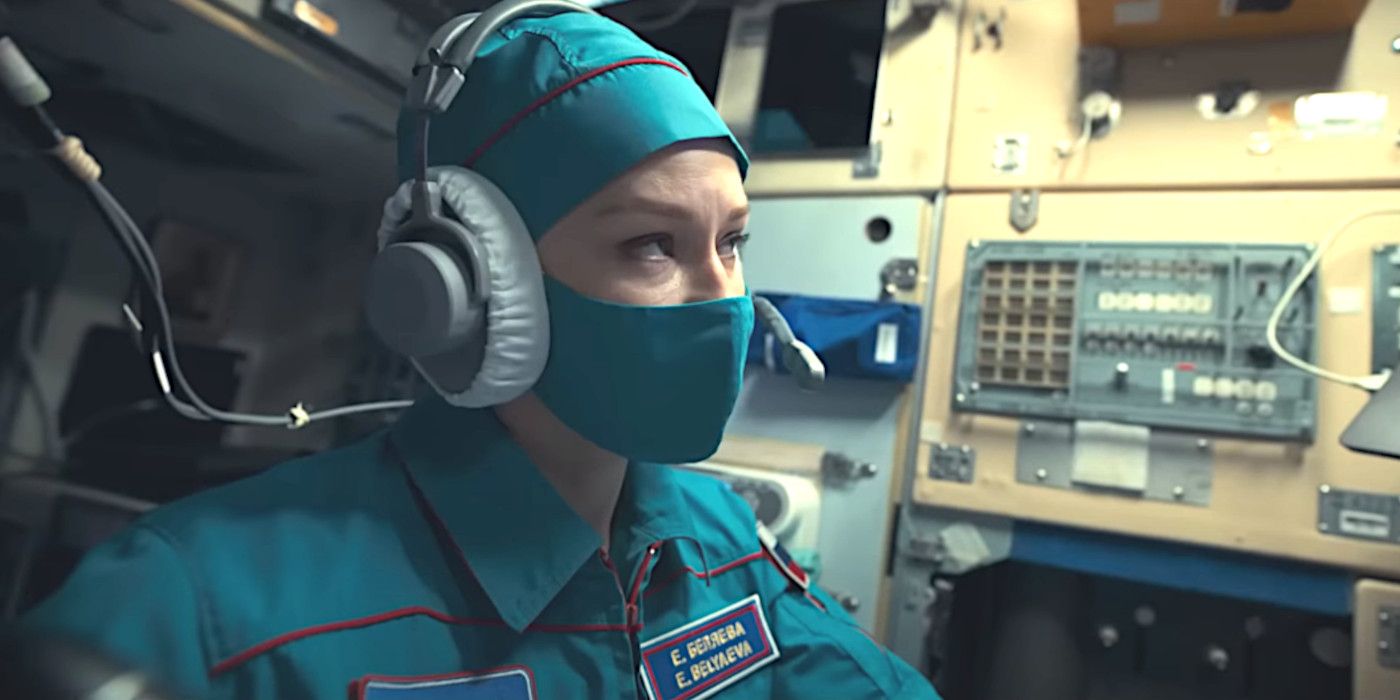 Uma mulher vestindo uniforme de cosmonauta azul, máscara cirúrgica e fones de ouvido, cercada por equipamentos eletrônicos