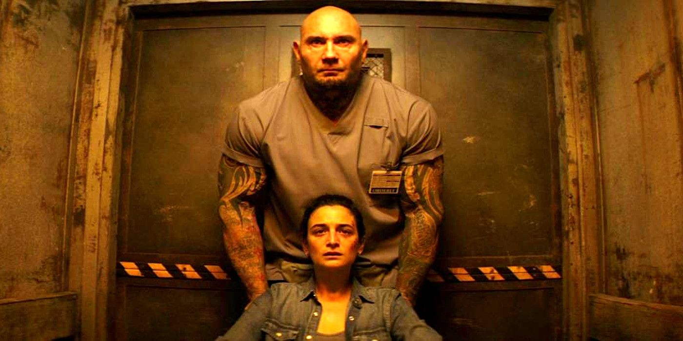 Dave Bautista in a freight elevator in scrubs, standing behind a wheelchair-bound Jenny Slate
