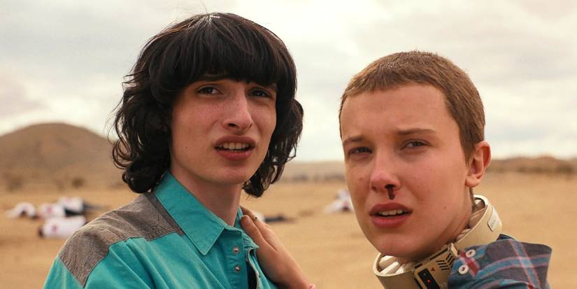 Eleven holds onto Mike while wearing a collar and having a nosebleed as they stand in an empty field looking on in Stranger Things