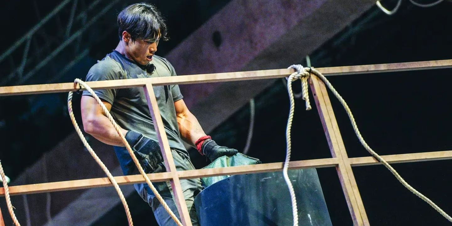 Physical 100 contestant Yun Sung-bin pouring a bag of sand into a black cylinder. He is wearing an all-black outfit with short sleeves and gloves. 