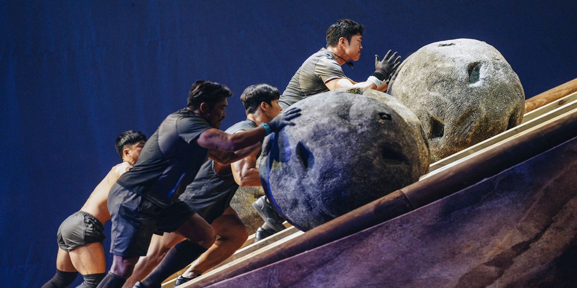 4 contestants on Netflix's Physical 100. They are all rolling large boulders up an incline. 