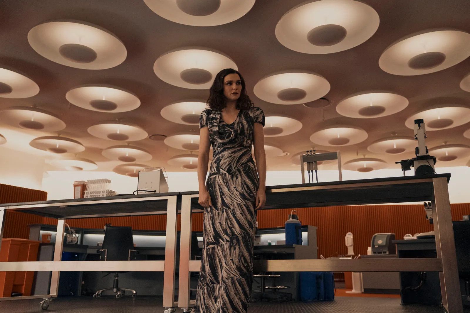 Rachel Weisz in Dead Ringers in a bold print dress standing against a table in a retro-futurist room