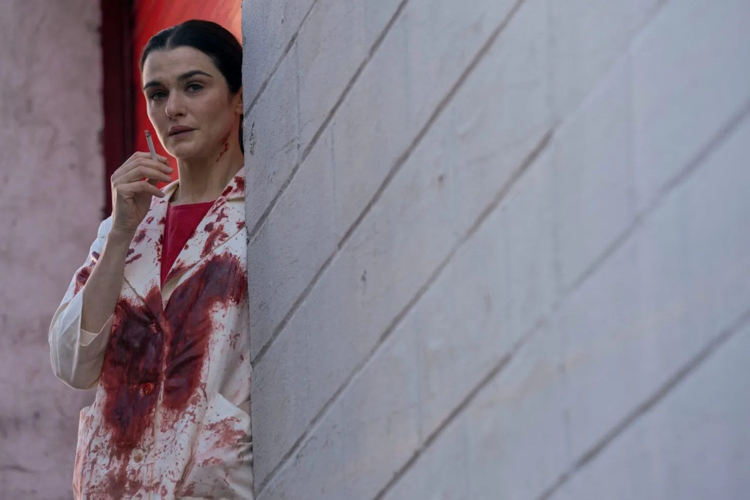 Rachel Weisz in a blood-soaked doctor's coat standing against a brick wall smoking a cigarette