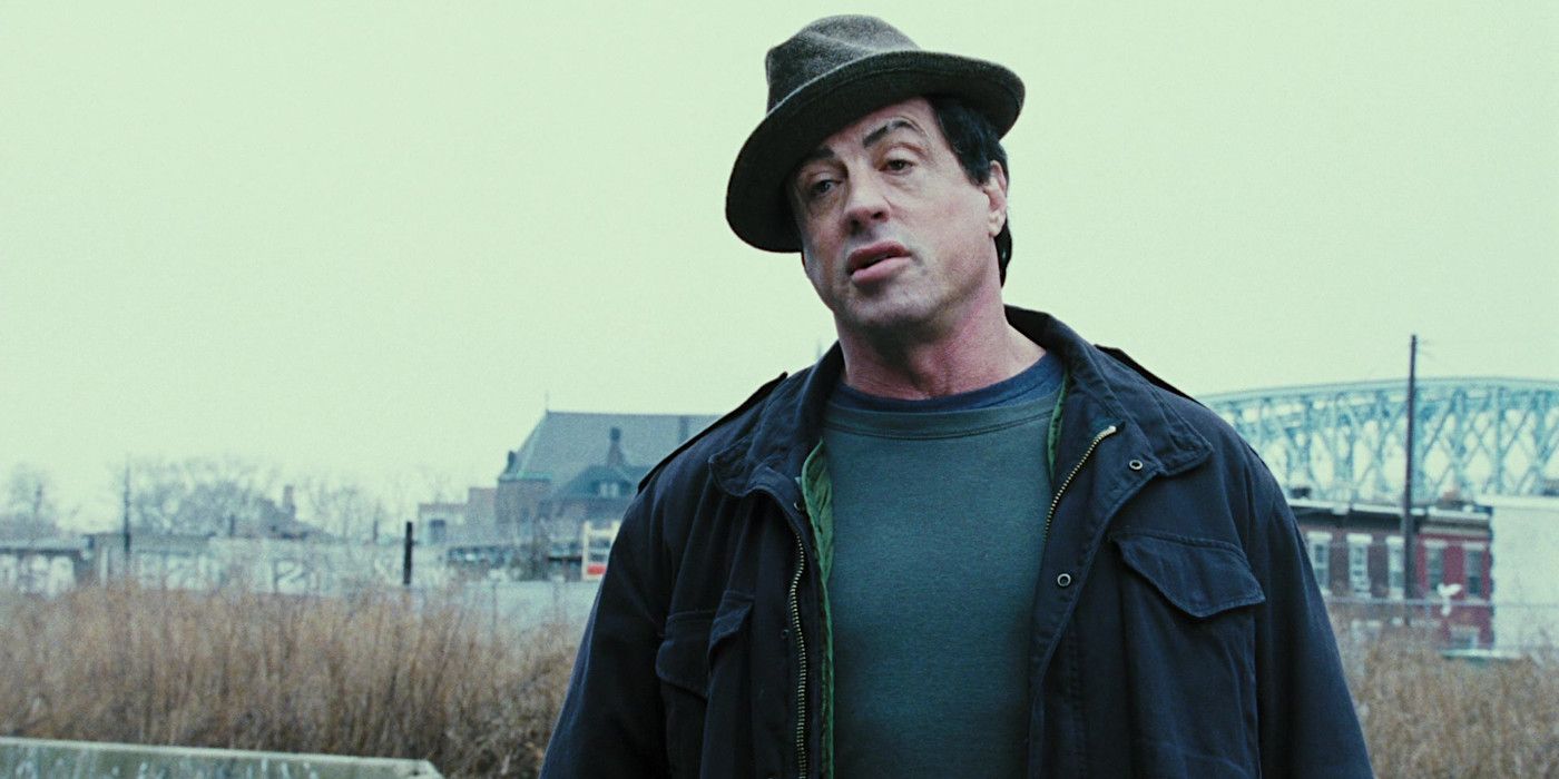 Sylvester Stallone in Rocky Balboa jacket and fedora in a cold exterior with industrial buildings and curly brush in the background