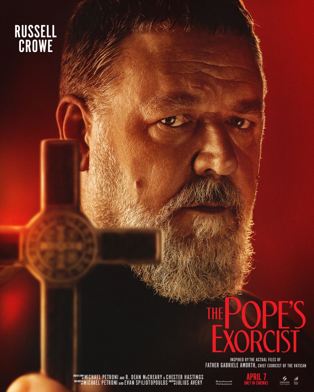 Russell Crowe poses dramatically in front of a devilish red background while holding up a wooden crucifix in The Pope's Exorcist movie poster