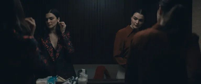 Rachel Weisz as twins in Dead Ringers, both standing in front of a mirror getting ready to go out, one dressed in a black outfit adjusting her earring, the other wearing a red buttoned-down shirt