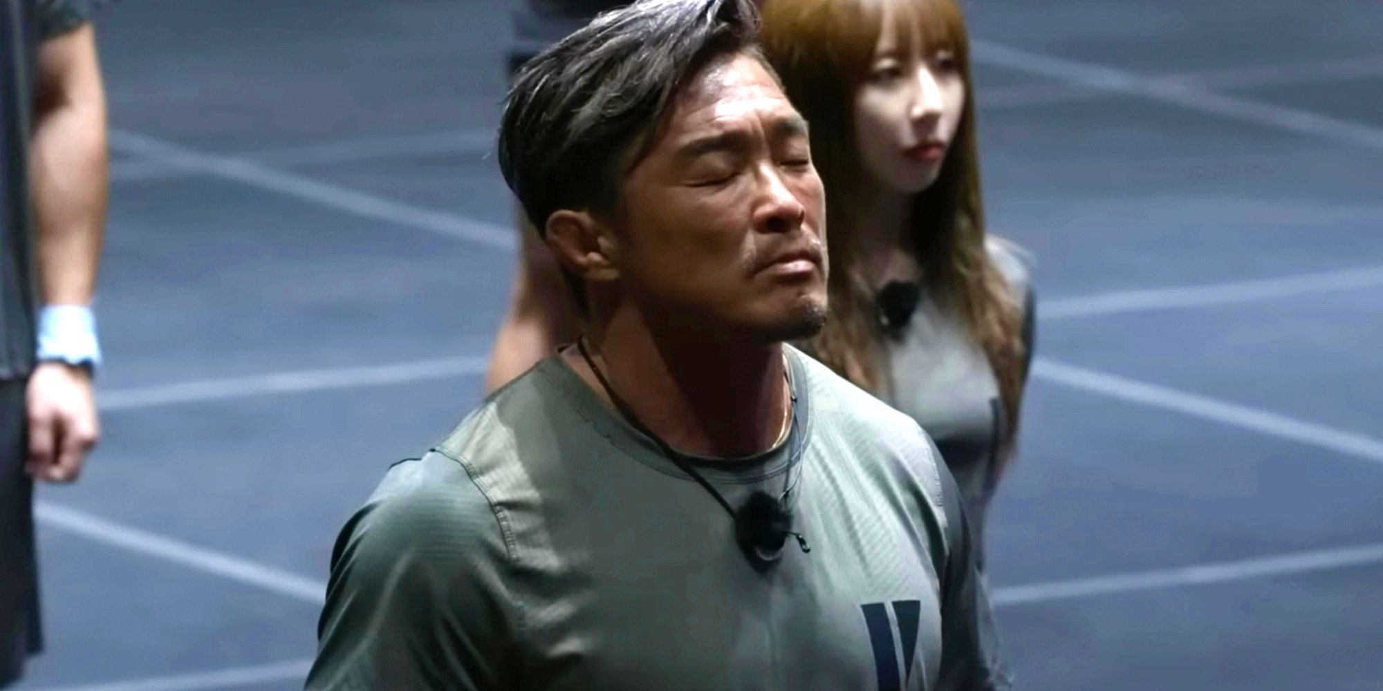 Physical 100 contestant Choo Sung-hoon. He is standing with his eyes closed in front of a few other competitors, wearing a gray tee shirt with a mic hanging around his neck. 