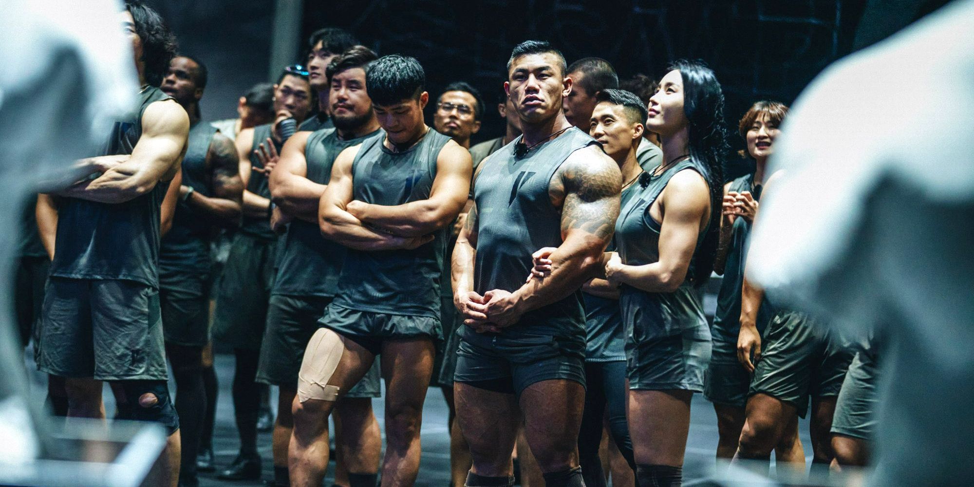 A line of Physical 100 contestants stand shoulder to shoulder, looking off in different directions. They are all wearing matching all-black workout clothes.