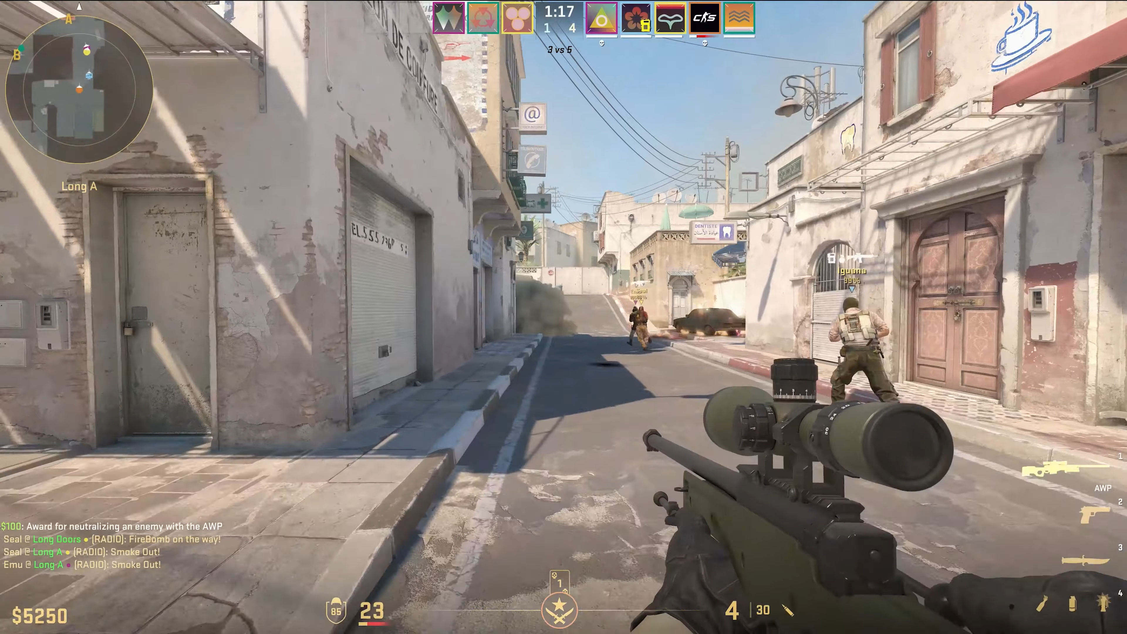 An in-game screenshot of Counter-Strike 2, showing the player walking through a city street carrying a sniper rifle with two other players ahead of them.