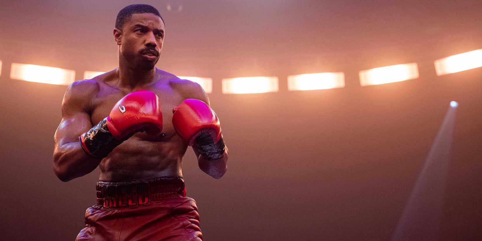 Adonis stands in the ring with his gloves up and bright spotlights shining behind him in Creed III