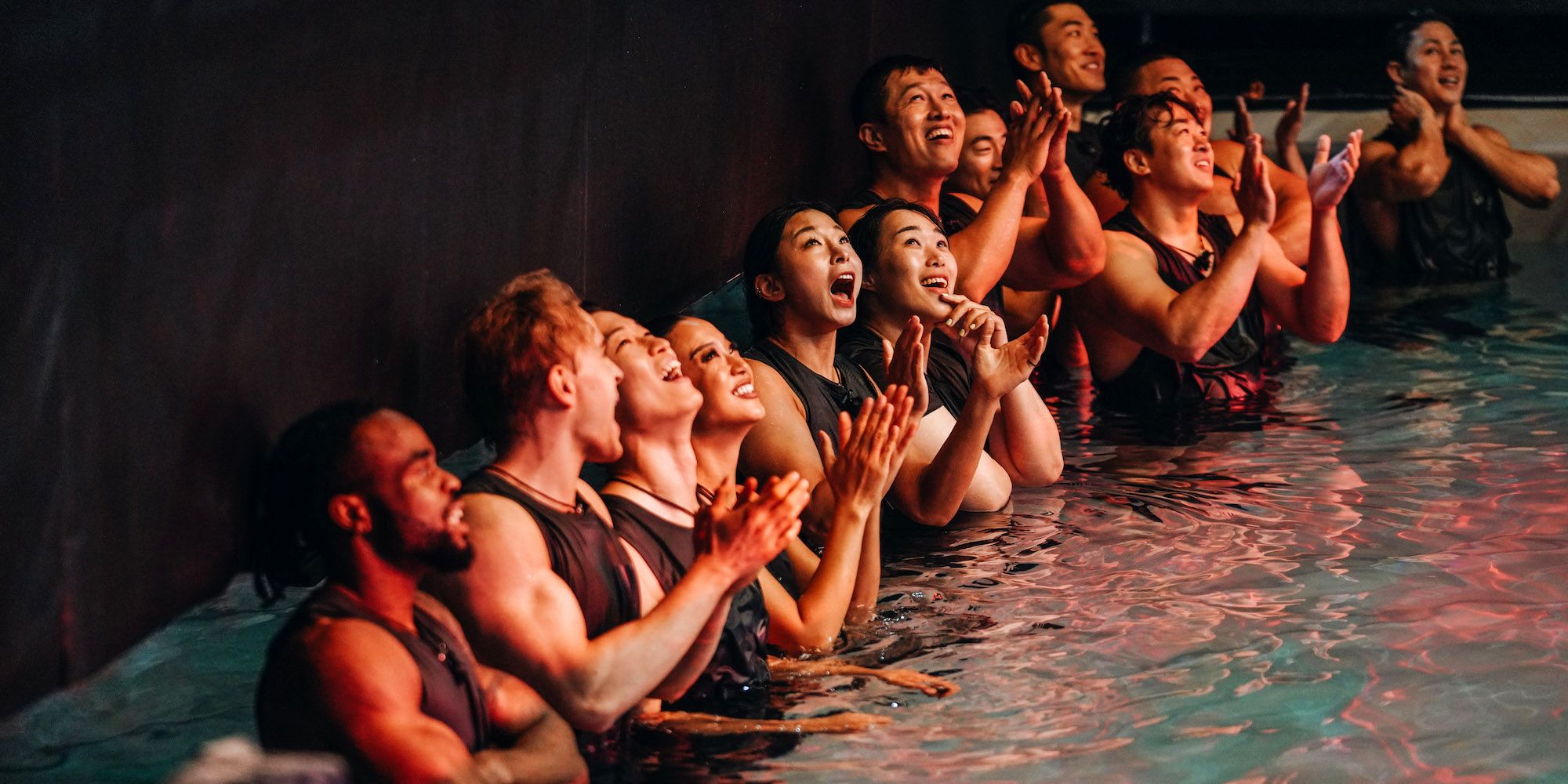 100 competidores físicos alinhados lado a lado em uma piscina. A água chega até a cintura e todos estão olhando para cima, torcendo pelos adversários.