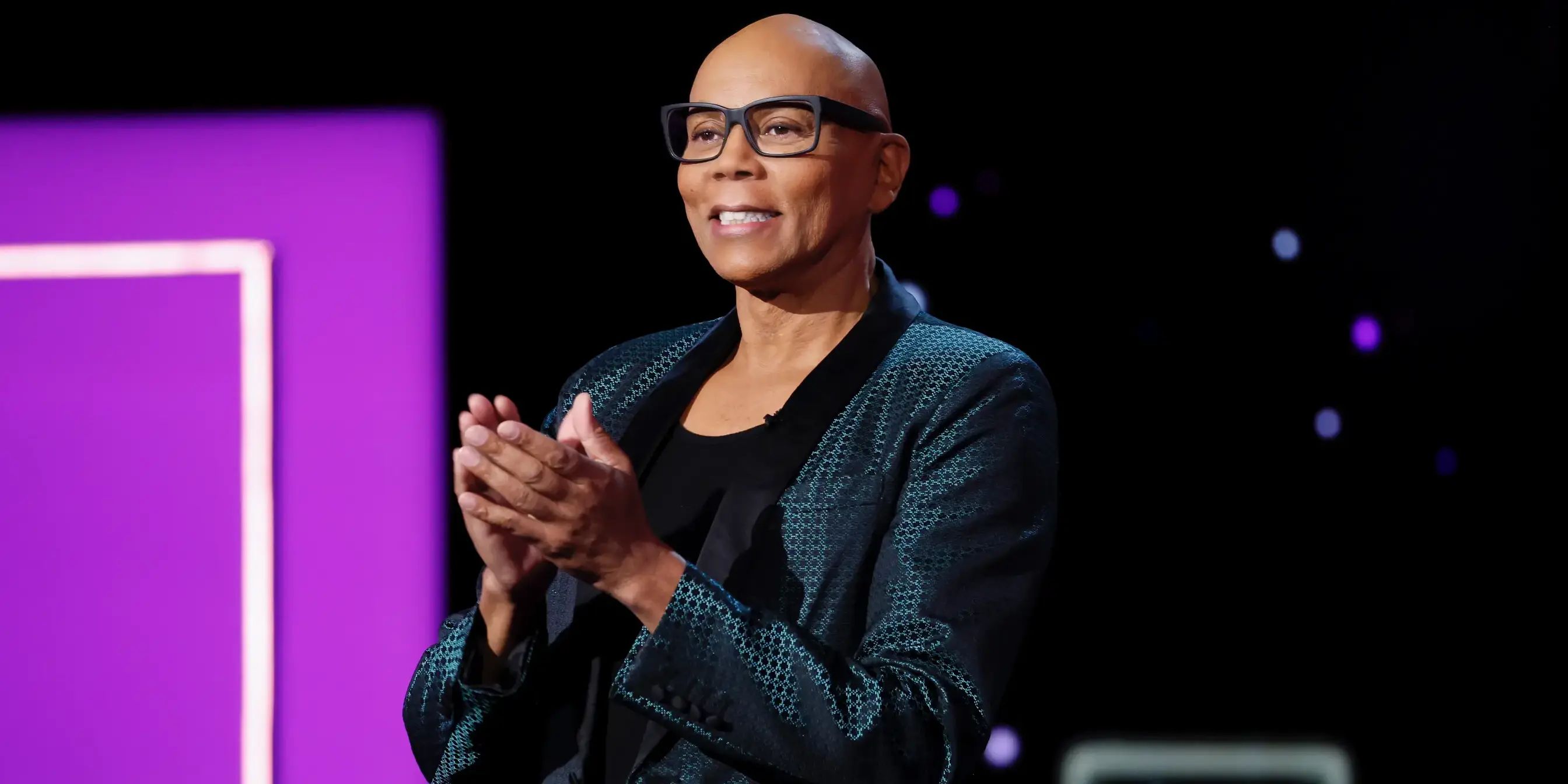 RuPaul from RuPaul's Drag Race smiles and claps in a navy blue suit