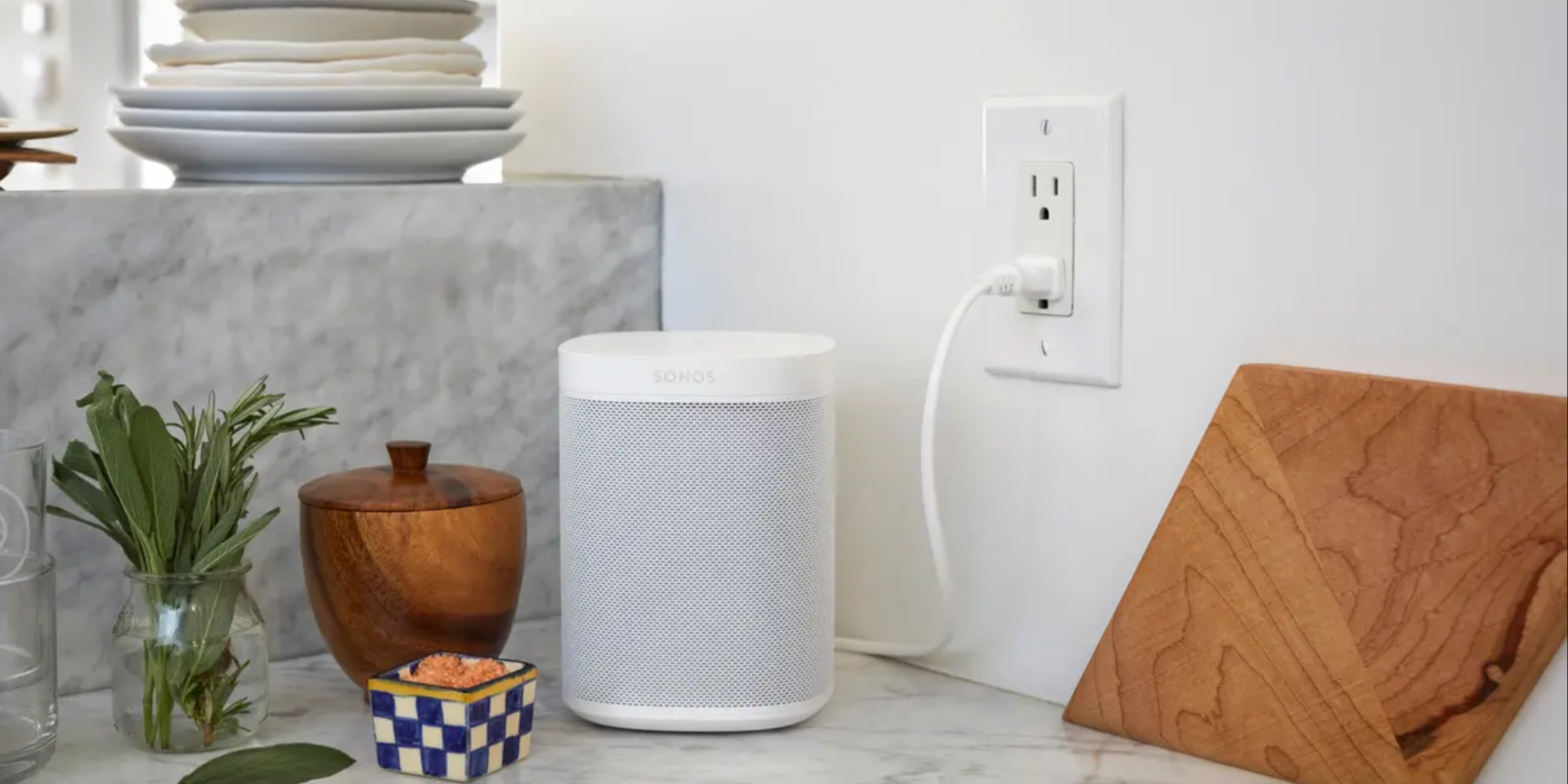 A white Sonos One speaker is pictured on a marble counter plugged into the wall beside a wooden bowl, herbs, and cutting board