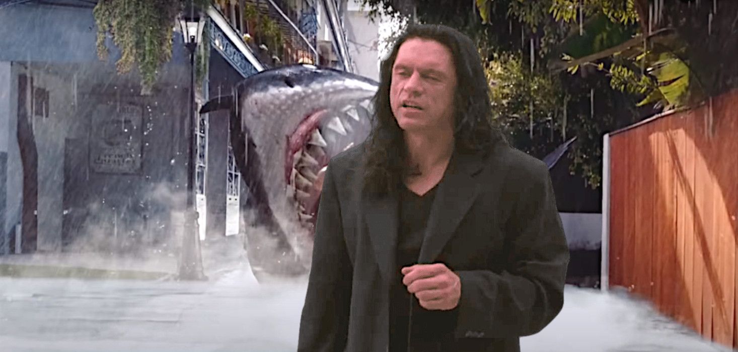 Tommy Wiseau in The Room in a black coat talking backdropped by a shark improbably attacking people on a New Orleans street