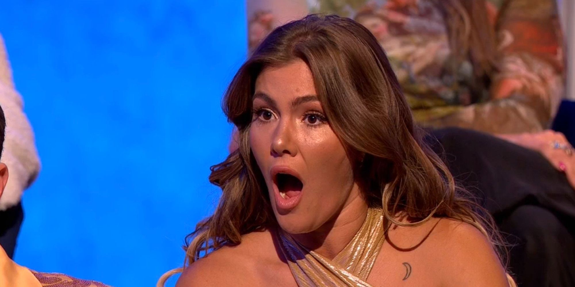 Tori Deal at the Challenge: Ride or Dies reunion. She is sitting against a blue background with her mouth open in shock and a surprised look on her face. Her long brown hair is down and she is wearing a gold top. 