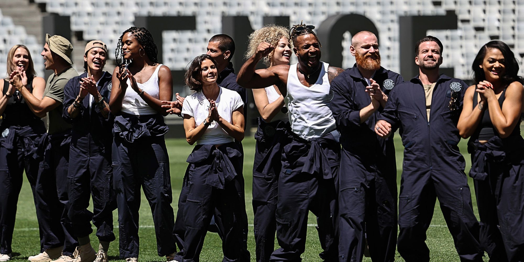 11 Challenge champions stand in a line on a football field. They are all wearing matching blue jumpsuits and cheering, posing, and smiling excitedly.