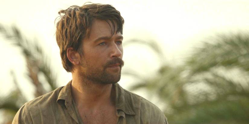 Brandon Sklenar as Spencer Dutton in 1923 season 1 with messy hair and mustache against backdrop of palm trees