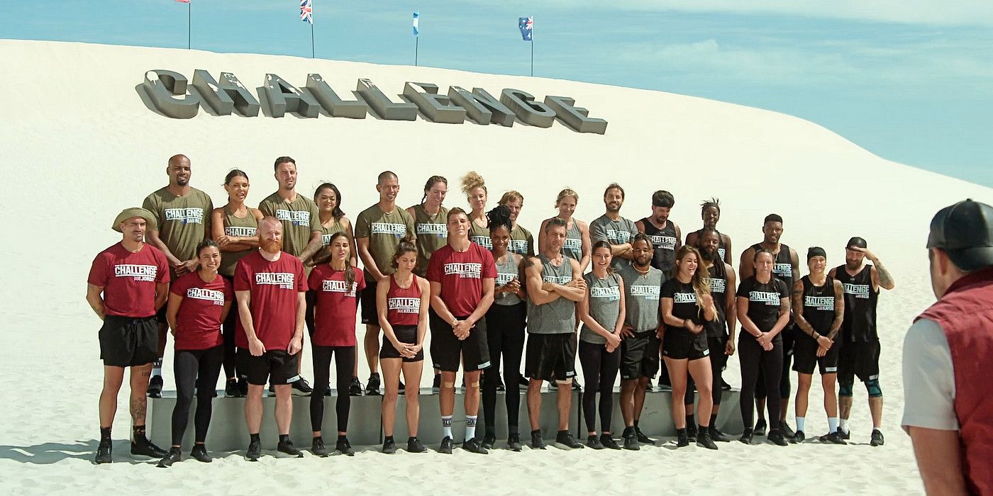 Contestants on The Challenge: World Championship standing side to side in two rows. They are wearing red, black, gray, and green shirts and standing against a backdrop of a white sand dune with The Challenge logo spelled out in giant letters. 