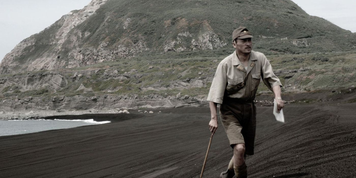 Screencap of Ken Watanabe as General Tadamichi Kuribayashi in Letters from Iwo Jima. Watanabe walks with a cane along the black sands of the island, with Mount Suribachi in the background.