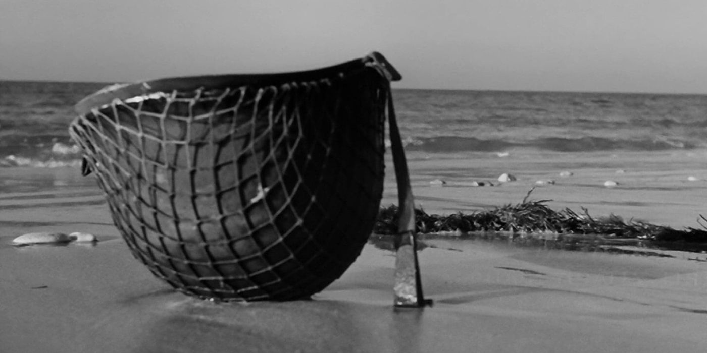 Screencap from The Longest Day (1962). A US army helmet with netting placed upside down on the sands of Normandy, with the English Channel in the distance.