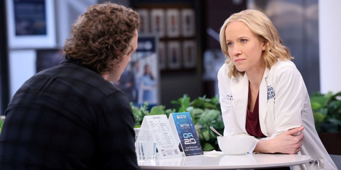 Hannah Asher and Sean Archer sit at a table in the hospital in Chicago Med season 8, episode 20. Asher wears a white doctor's coat over maroon scrubs. Sean wears a dark blue plaid shirt.