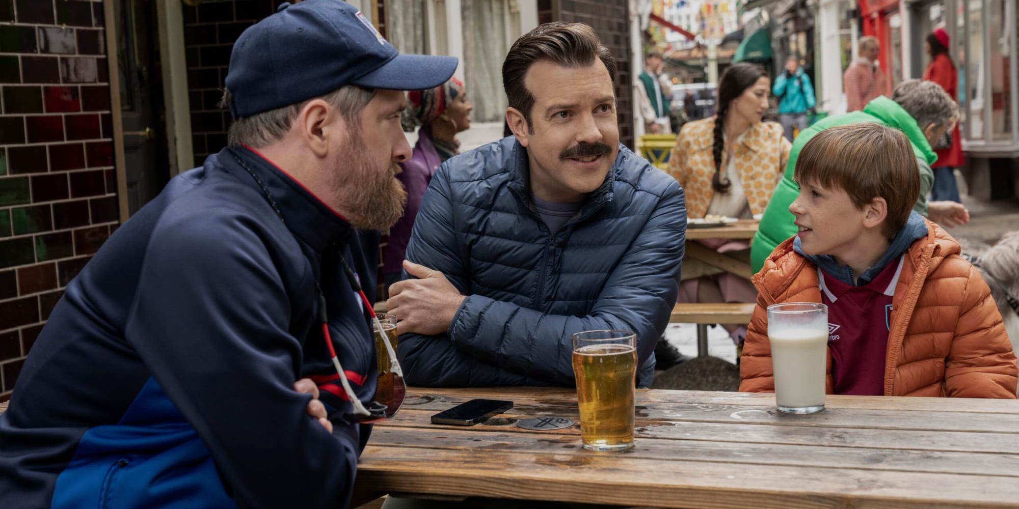 Coach Beard (Brendan Hunt), Ted (Jason Sudeikis), and Henry (Gus Turner) on a pub terrace in Ted Lasso season 3