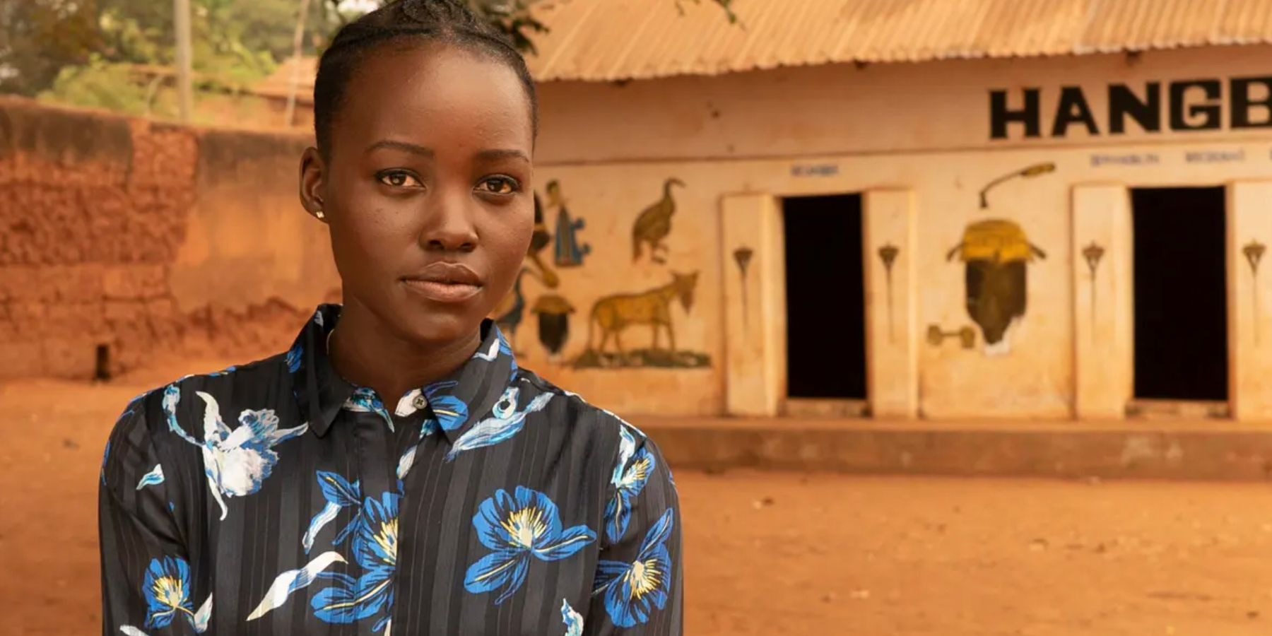 Lupita Nyong'o on an orange clay road and in front of a white building in Warrior Women