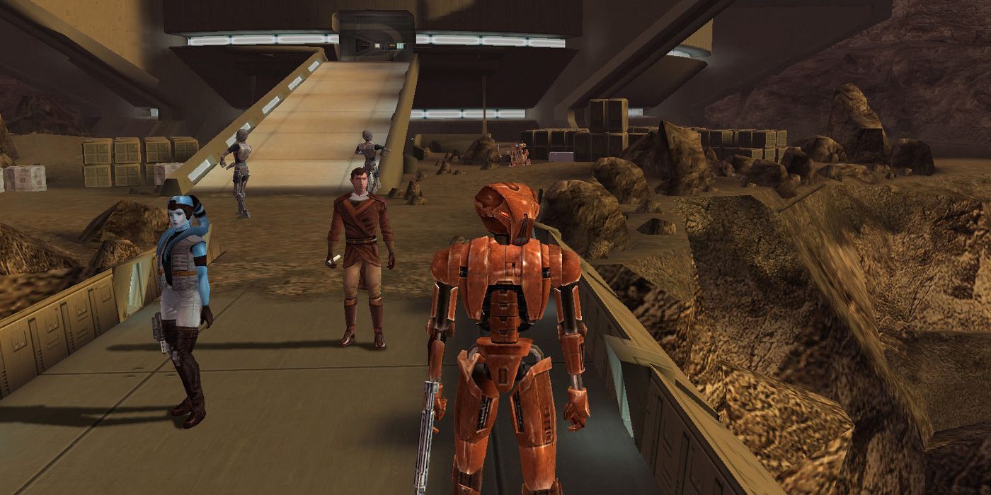 A player character wearing bronze-colored armor and carrying a blaster stands in a crowded walkway in front of a large, futuristic building in Star Wars: Knights of the Old Republic.
