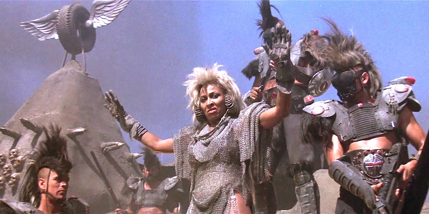 Tina Turner as Auntie Entity in Mad Max Beyond Thunderdome wearing chain mail and huge looping metallic earrings, gesturing for everyone to quiet down while addressing a crew of post-apocalyptic warriors decked out in improvised armor