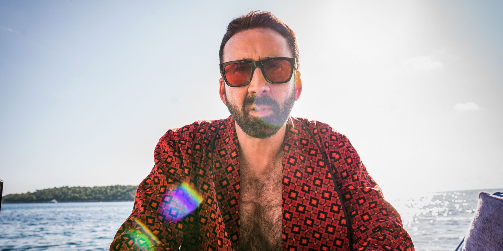 Nicolas Cage, wearing a luxurious red robe and a big pair of sunglasses, sits waterside in The Unbearable Weight of Massive Talent.