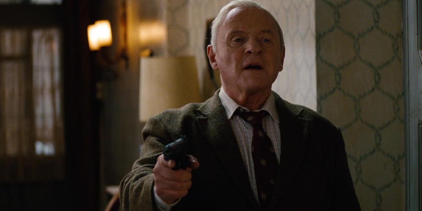 Bailey from Red 2 holds a gun up while wearing a suit in a dimly lit hallway.
