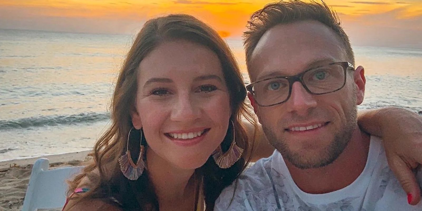 Danielle & Adam Busby smiling at the beach with the ocean and sunset behind them