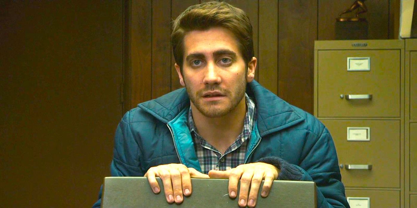 Jake Gyllenhaal en Zodiac luciendo impecable con una chaqueta azul, sentado en una silla en una oficina, luciendo decepcionado