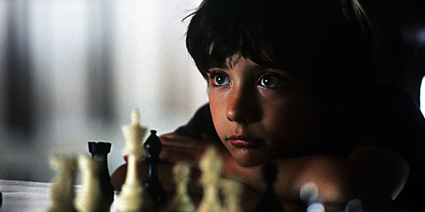 Josh leans his arms and chin on a chess board, looking away, with chess pieces in front of him.