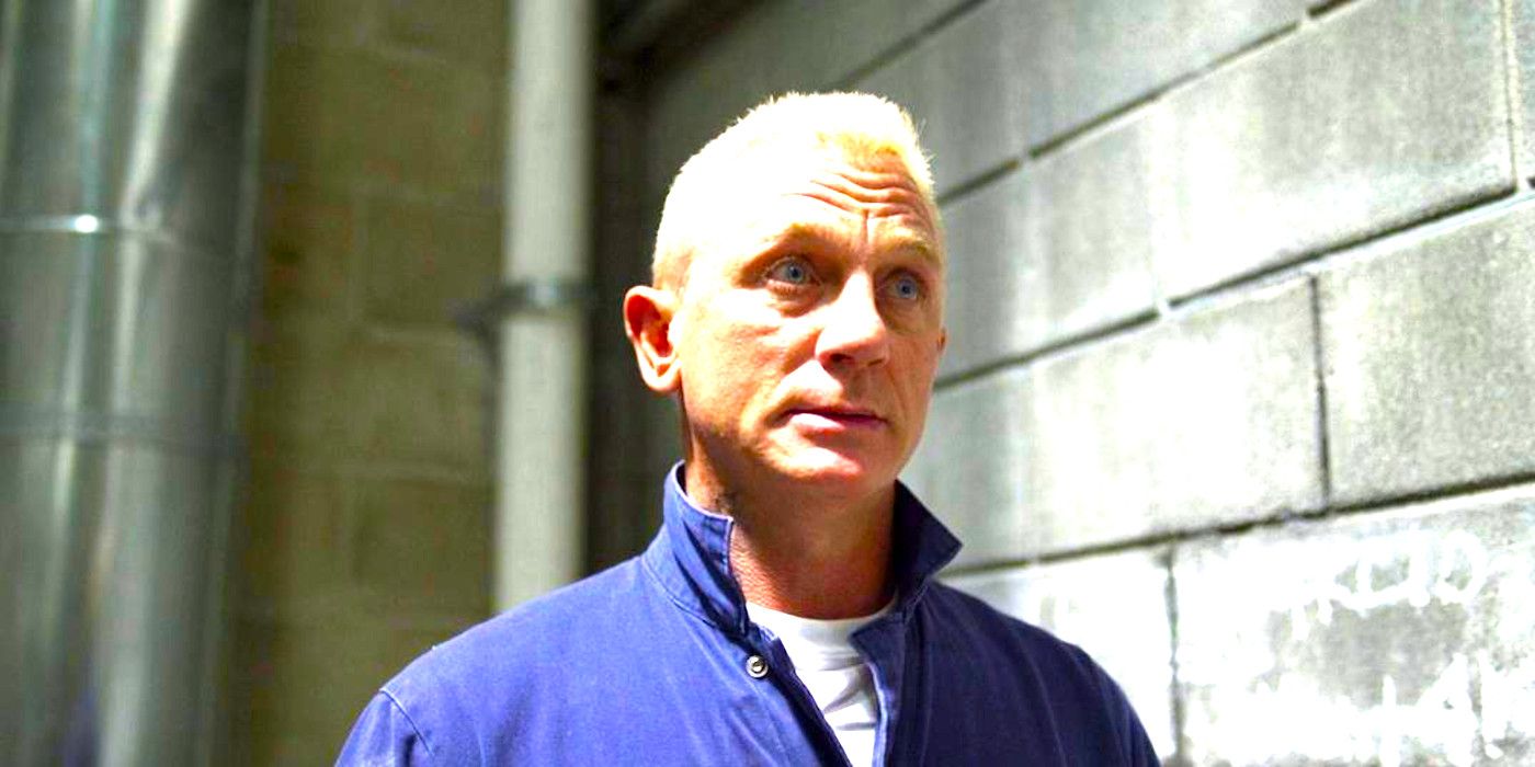 Daniel Craig in Logan Lucky with bleached blond hair and crazy eyes, wearing a blue jumpsuit against a cinderblock wall