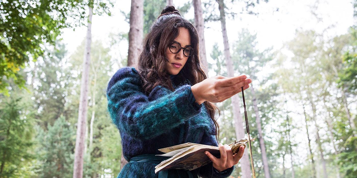 Anathema holds up a talisman while reading a book in the woods in Good Omens